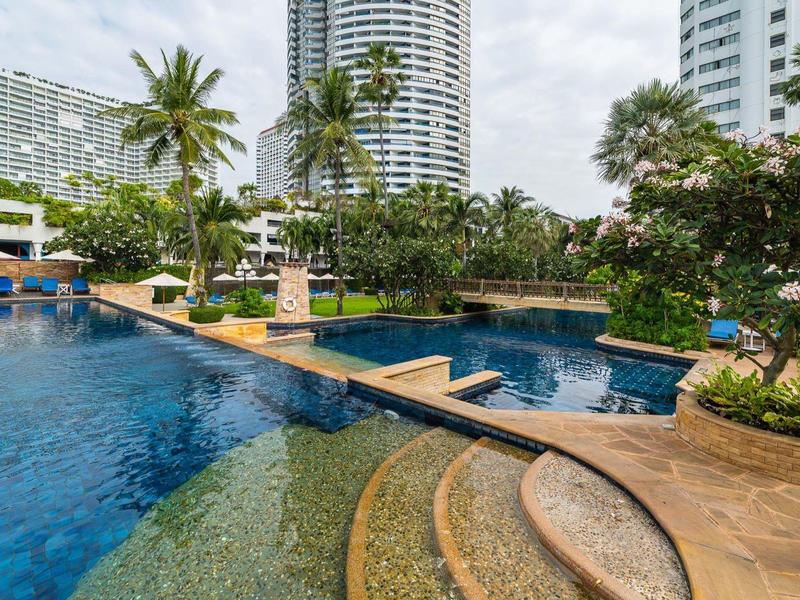 Großer Außenpool mit Treppen, Palmen und Hochhäusern im Hintergrund unter bewölktem Himmel.