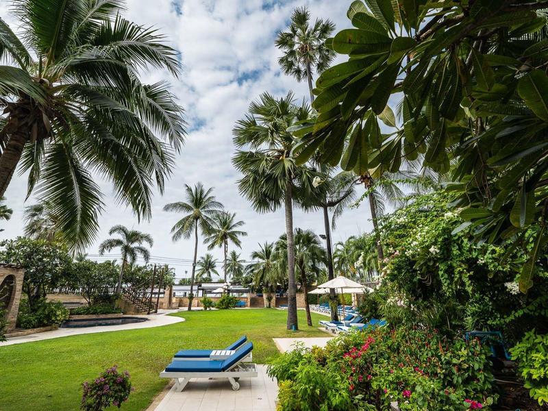 Grüner Garten mit hohen Palmen, blauem Liegestuhl und weißem Sonnenschirm unter bewölktem Himmel.