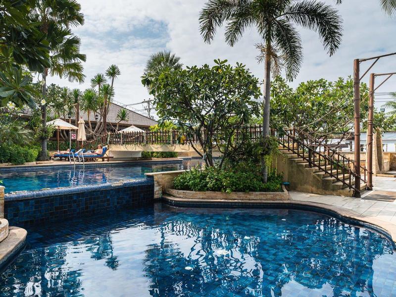 Blauer Pool mit Wasserfall, umgeben von Palmen, Bäumen und einem Holzsteg unter bewölktem Himmel.