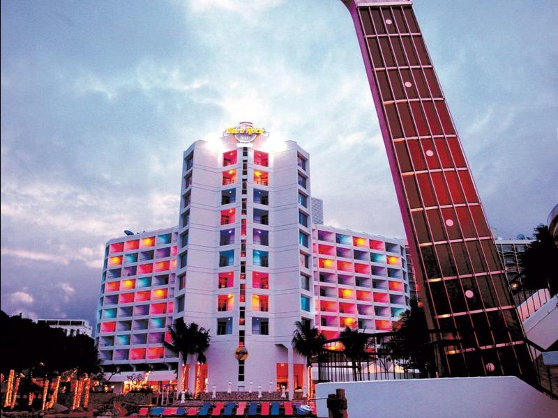 Bunte Hotelbeleuchtung spiegelt sich im Pool, davor großer Gitarrenhals in Abenddämmerung.
