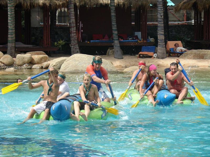 Sechs Personen paddeln auf blauen Luftballons im klaren Wasser vor einem Strand mit Palmen.