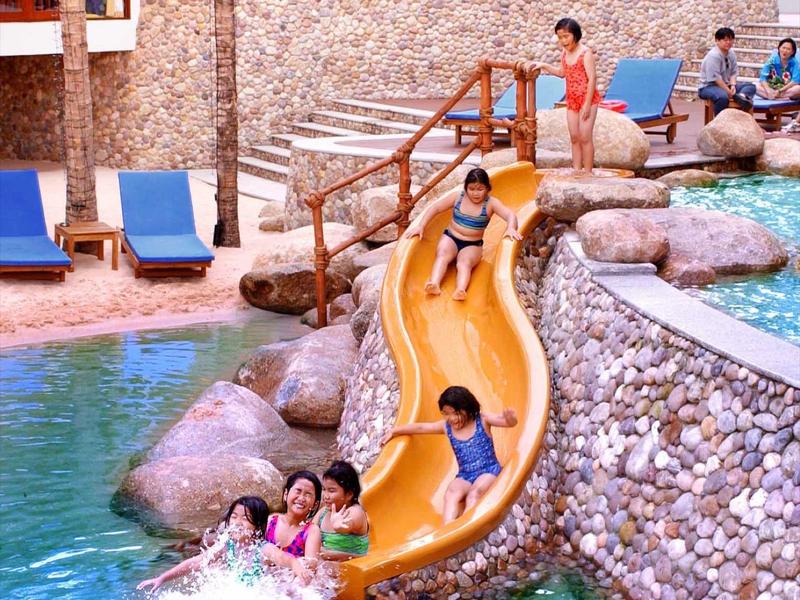 Kinder rutschen eine gelbe Rutsche in ein Pool mit steinverkleideter Umrandung und blauen Liegestühlen.