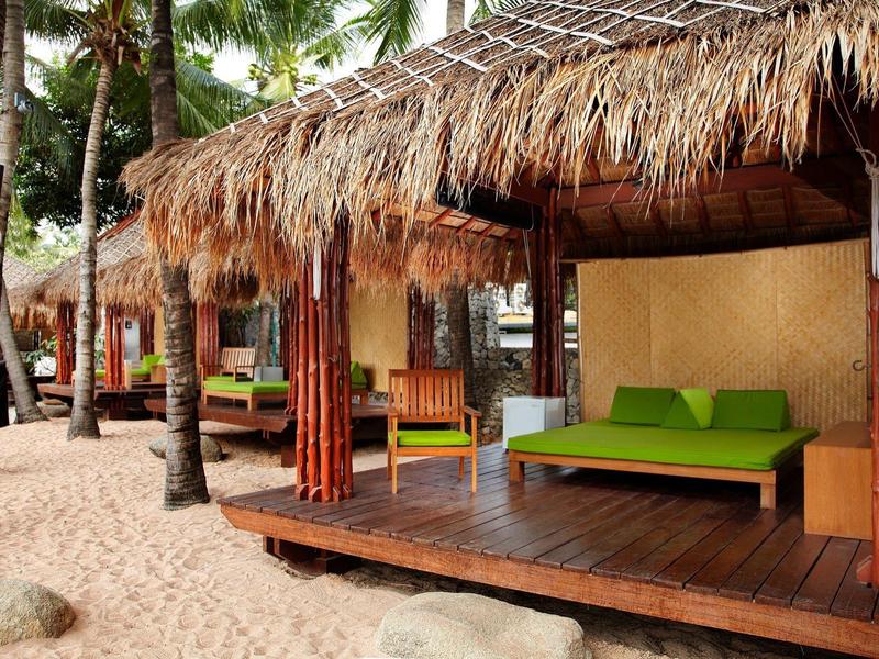 Strandbungalows mit Strohdächern, Holzterrassen und grünen Sitzkissen umgeben von Palmen.