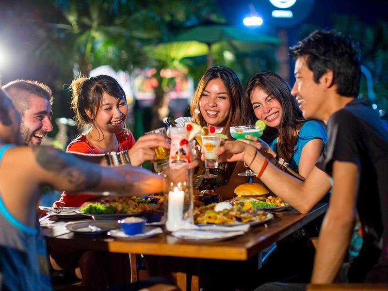 Fünf Freunde stoßen lachend mit Getränken an, sitzen draußen an einem beleuchteten Holztisch.