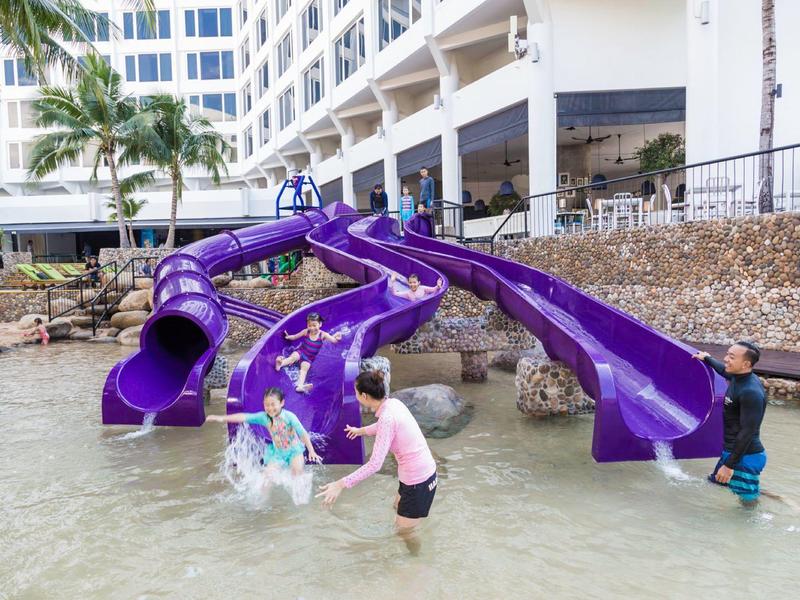 Kinder spielen in flachem Wasser an zwei großen, geschwungenen violetten Wasserrutschen neben einem Hotel.