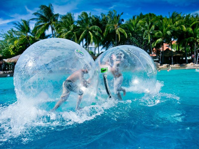 Zwei Personen in großen, durchsichtigen Wasserbällen rennen auf einem Pool mit blauem Wasser.