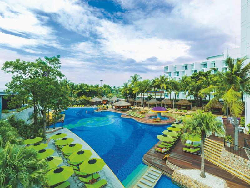 Großer blauer Pool mit Liegestühlen, Palmen und weißem Hotelgebäude unter blauem Himmel.
