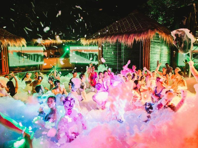 Menschen tanzen auf einer Schaum-Party im Freien bei bunten Lichtern und Strohdachhütten.