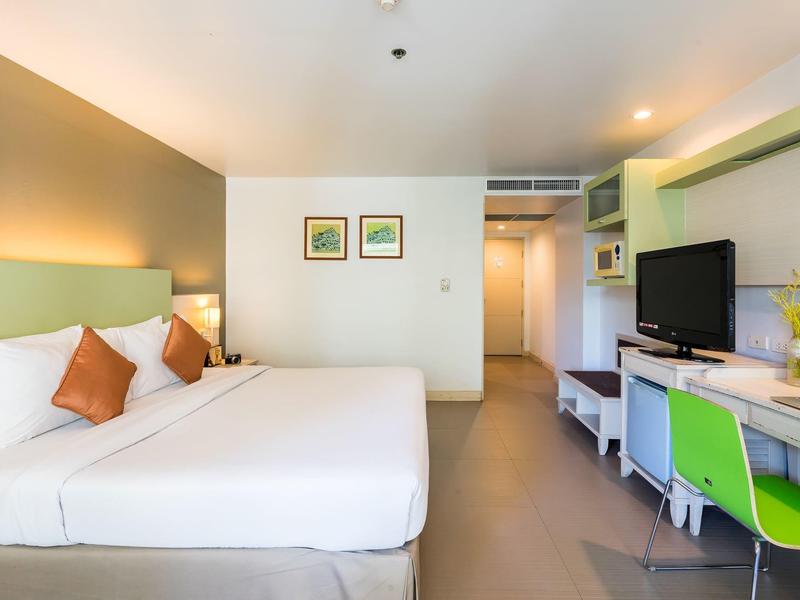 Hell eingerichtetes Hotelzimmer mit zwei Betten, grünem Stuhl und Flachbildfernseher auf Schreibtisch.