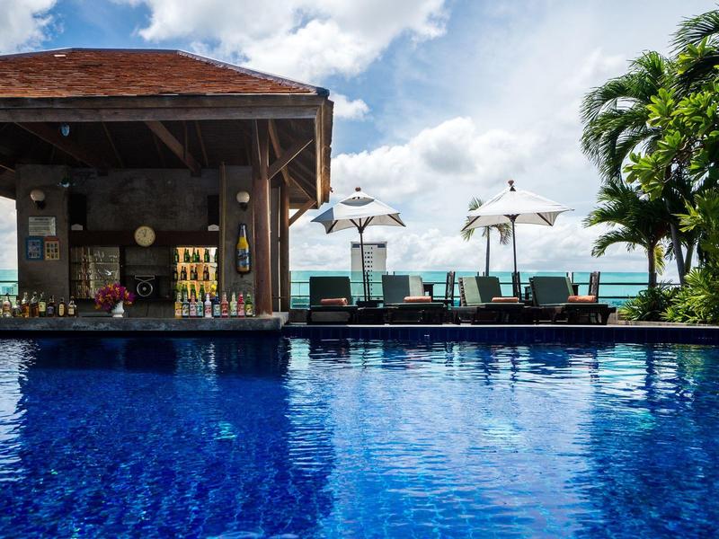 Freizeitbereich mit blauem Pool, Holzhaus, Liegestühlen und Palmen unter hellem Himmel.