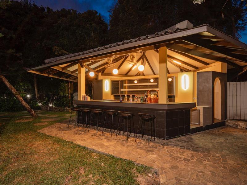 Beleuchteter Gartenpavillon mit Holzbar und Sitzplätzen bei Dämmerung, umgeben von Rasen und Bäumen.