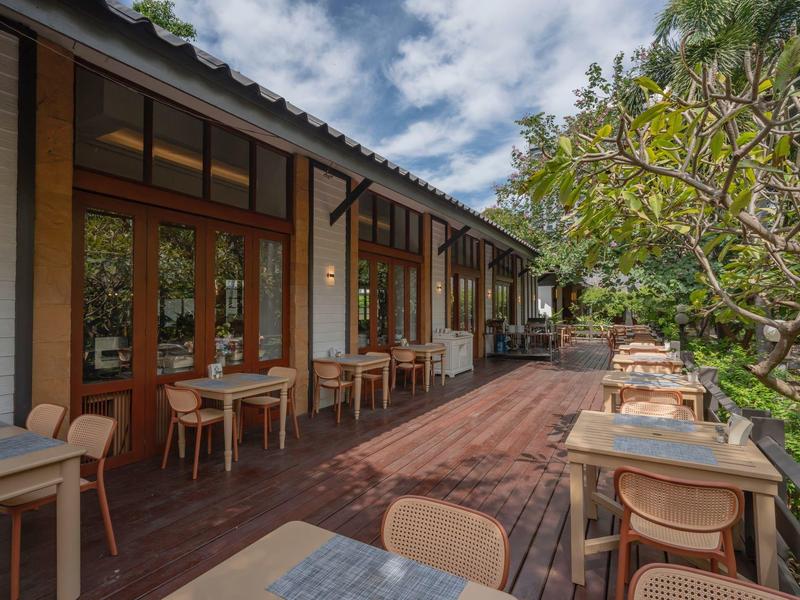 Terrassenrestaurant mit Holzboden, Tischen und Stühlen, umgeben von grünem Baum und bewölktem Himmel.