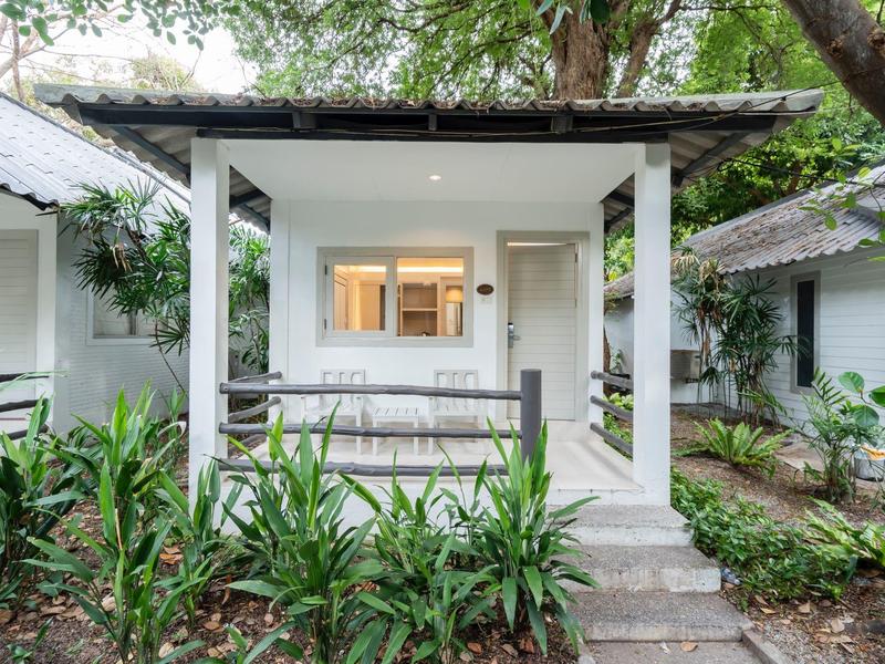 Kleines weißes Haus mit Veranda, umgeben von grünen Pflanzen und Bäumen, Eingangstür und Fenster sichtbar.