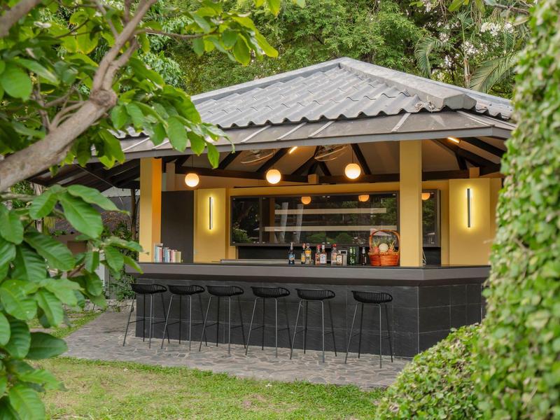 Offene Gartenbar mit dunklem Tresen und Hockern, umgeben von grünem Laub und Bäumen.