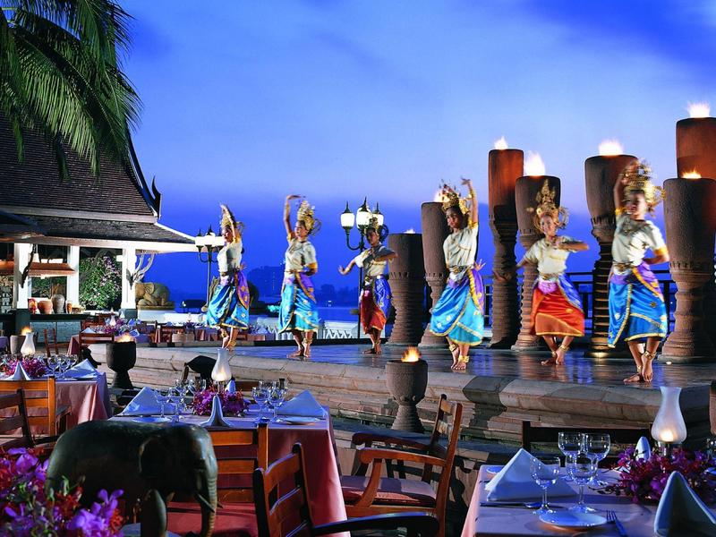 Tänzer in bunten Kostümen führen traditionelle Aufführung vor blauem Himmel auf Terrasse mit Esstischen auf.