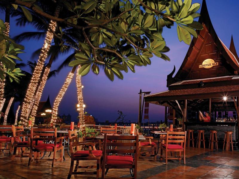 Freiluftrestaurant mit Holztischen, Stühlen, Palmen und traditionellem, spitzem Dach bei Dämmerung.