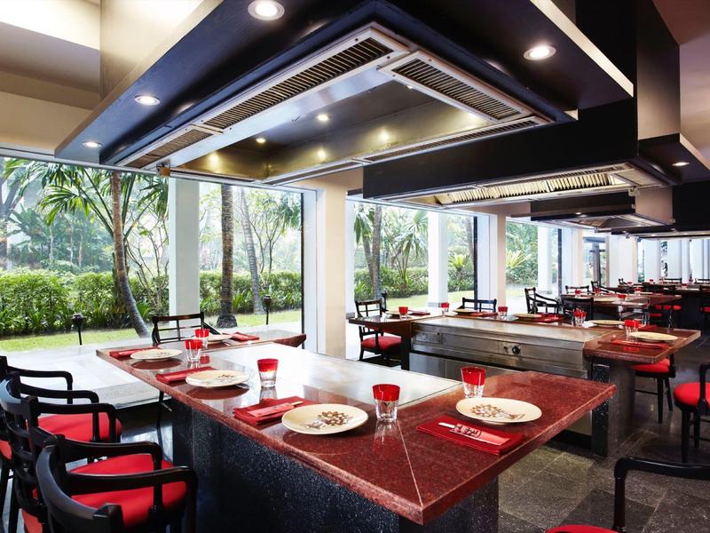 Modernes Restaurant mit großen Fenstern und roten Stuhlpolstern, verbunden mit grüner Außenanlage.