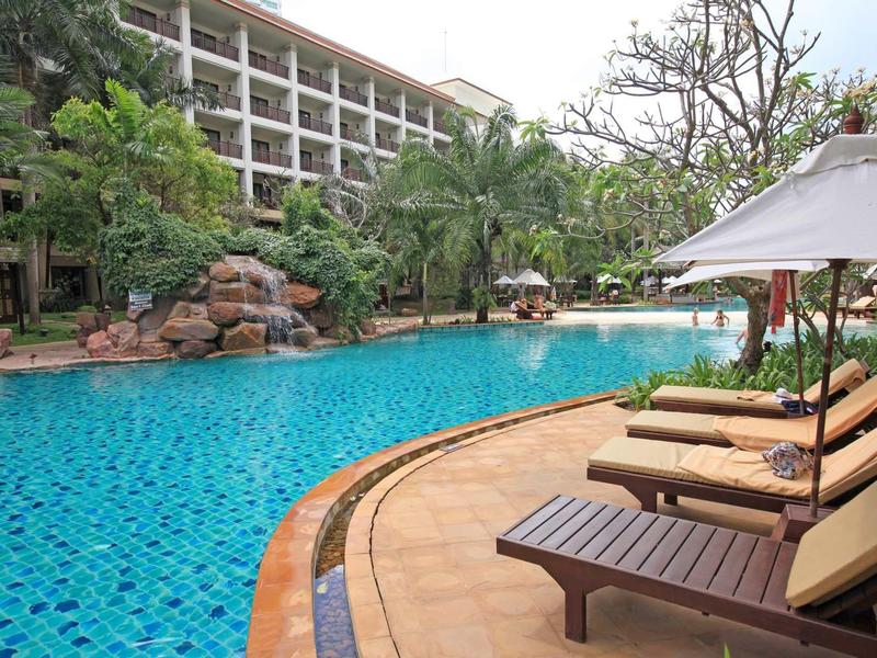 Großer blauer Pool neben Liegestühlen mit Schirmen und einem mehrstöckigen Hotelgebäude im Hintergrund.