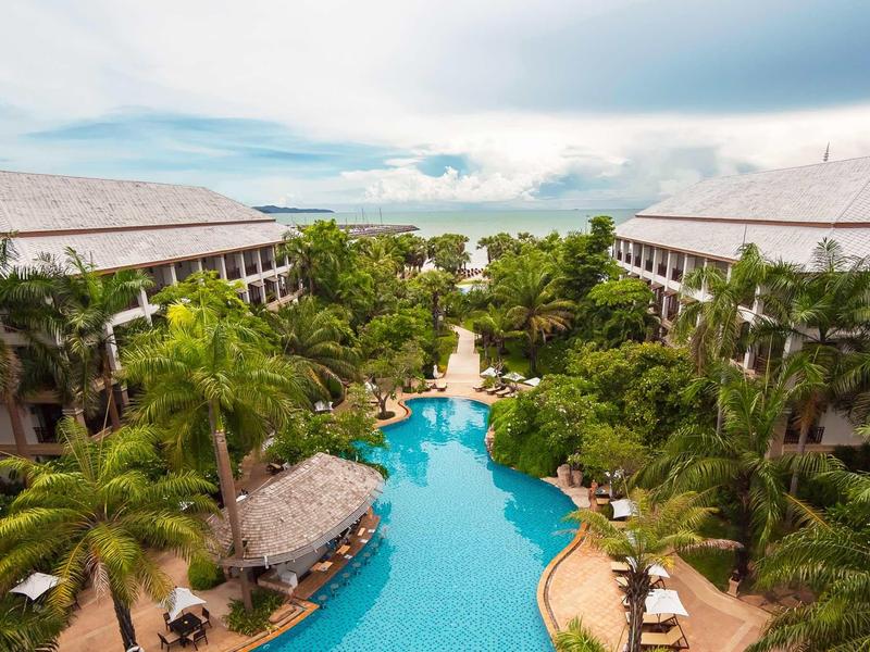 Blick auf einen tropischen Hotelpool mit Palmen, Liegestühlen und zwei großen Gebäuden.