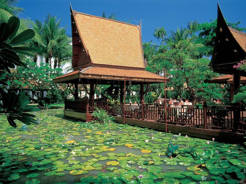 Traditionelle thailändische Holzhäuser mit spitzen Dächern über einem Teich mit grünen Seerosen.