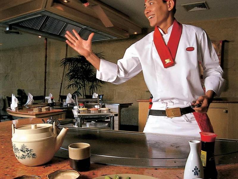 Koch in roter Mütze zeigt Sushi auf Teller mit Sojasauce und Essstäbchen an traditionellem Tisch.