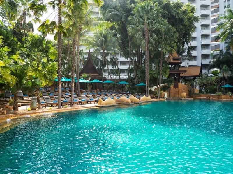 Großer blauer Pool umgeben von Palmen, Liegestühlen und Hochhäusern im Hintergrund.