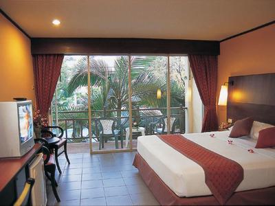 Hotelzimmer mit großem Bett, rotem Bezug, Glasfenster mit Blick auf Palmen und Fernseher auf Schreibtisch.