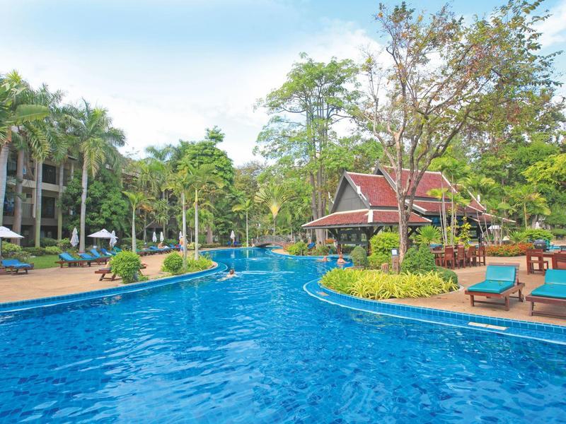 Großer blauer Pool mit Liegestühlen, grünen Palmen und Gebäuden unter bewölktem Himmel.