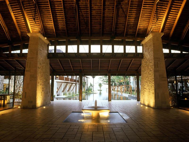 Großer Innenraum mit Holzdach, zwei beleuchteten Steinsäulen und hellem Eingang zum Außenbereich.