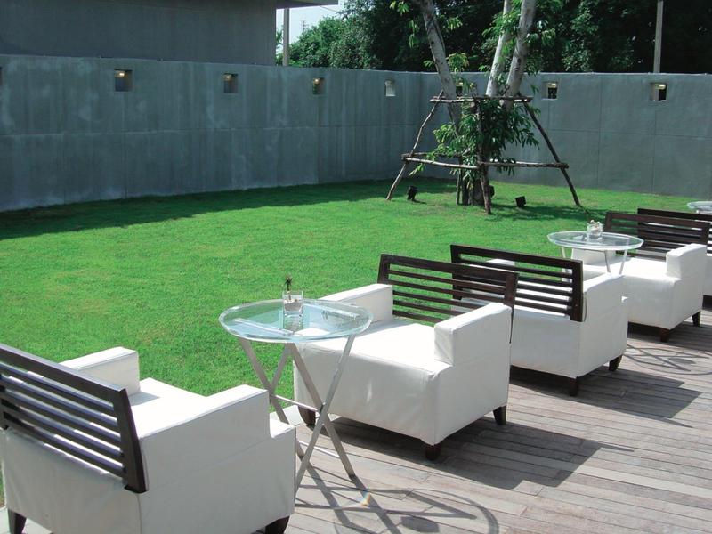 Weiße Sessel und Glastische stehen auf einer Terrasse neben einem grünen Rasen vor einer grauen Wand.