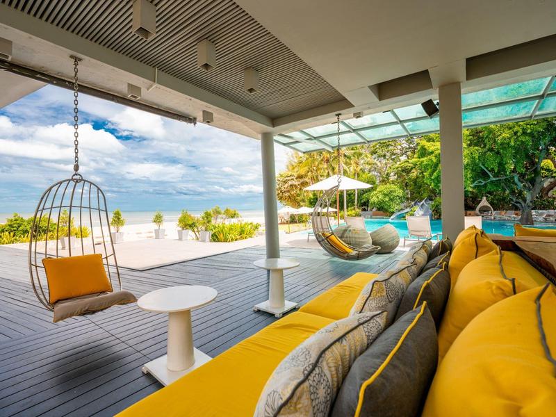 Offene Terrasse mit gelben Kissen, Hängesessel, Tisch und Blick auf Pool und Garten.