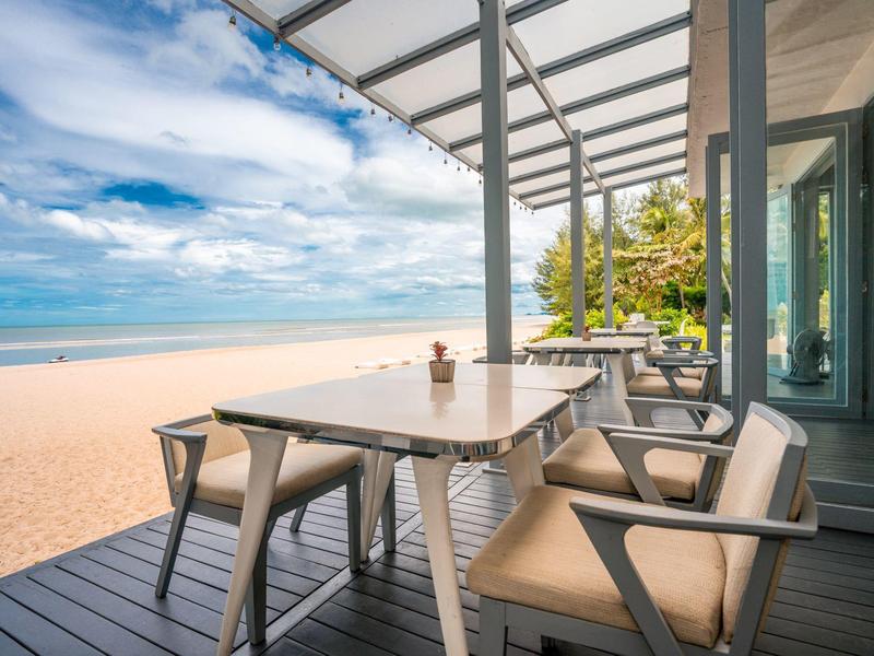 Terrasse mit Tisch und Stühlen am Strand, blauer Himmel und Meer im Hintergrund.