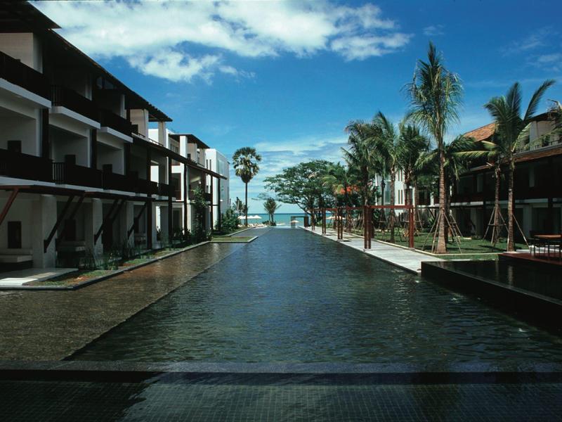 Ein langer, dunkler Pool zwischen weißen Gebäuden mit Palm Bäumen unter blauem Himmel.