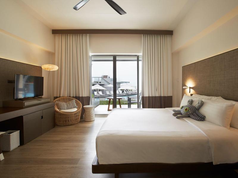 Modernes Hotelzimmer mit großem Bett, TV, Sessel und Balkon mit hellen Vorhängen und Holzfußboden.