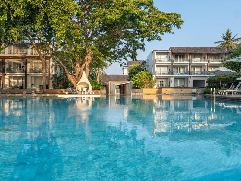 Großer blauer Pool vor modernen Hotelgebäuden mit Bäumen und Sonnenschirmen.