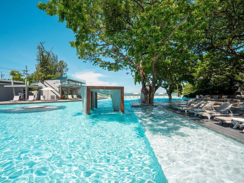 Moderner Außenpool mit Liegestühlen, Sonnenschirmen und großen Bäumen an sonnigem Tag.