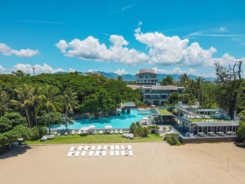 Strand mit Sand, weißer Liegen, großer Pool, Palmen und mehrstöckigem Gebäude im Hintergrund.