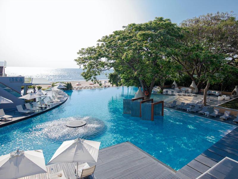 Einladender Pool mit Bäumen, Liegestühlen und Sonnenschirmen unter blauem Himmel am Meer.