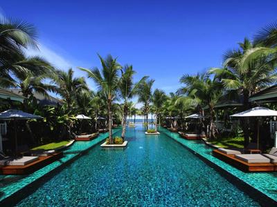 Long pool with palm trees and loungers under a bright blue sky in a tropical resort.