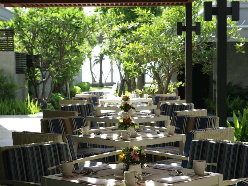 Restaurant terrace with tables and chairs under a sunshade in a green outdoor area.