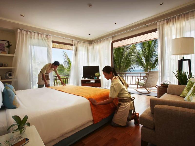 Two hotel staff preparing a bed in a bright, open room with a view of the terrace.