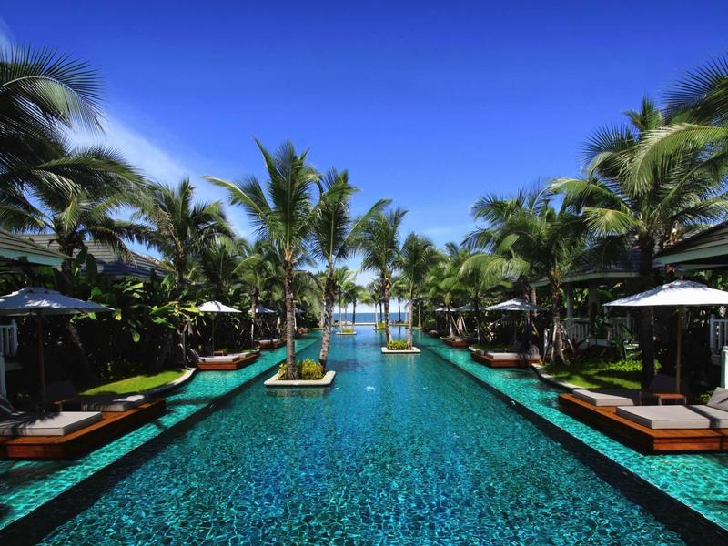 Long pool with palm trees and loungers under a bright blue sky in a tropical resort.