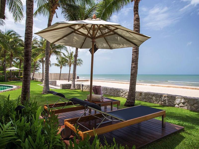 Beach loungers with umbrella under palm trees on grass next to a sandy beach and sea.