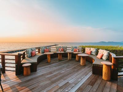 Holzterrasse mit gebogenem Sitzbereich und bunten Kissen mit Blick auf das Meer bei Sonnenuntergang.