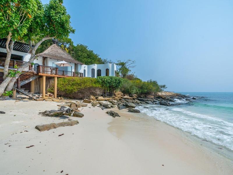 Beach with fine sand and rocks, adjacent buildings, green trees, and a blue sky.