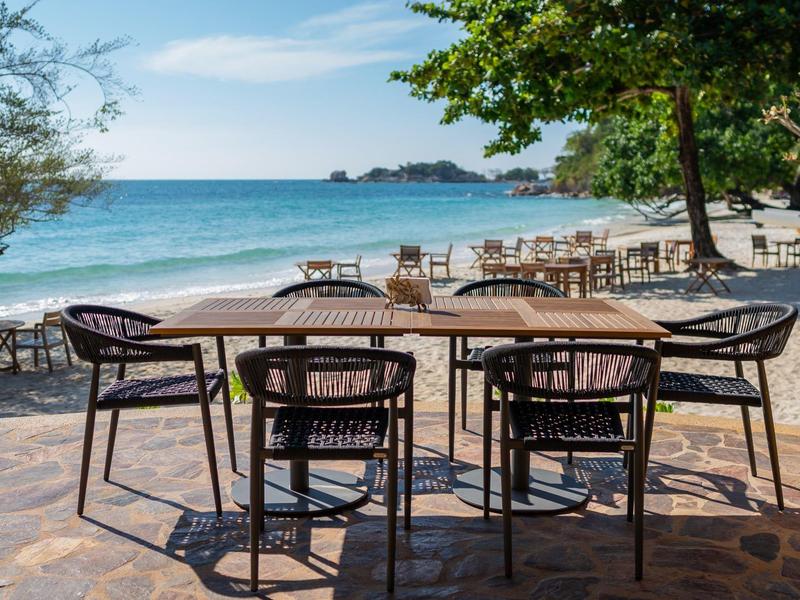Tables and chairs on a terrace by the beach overlooking the sea and trees.