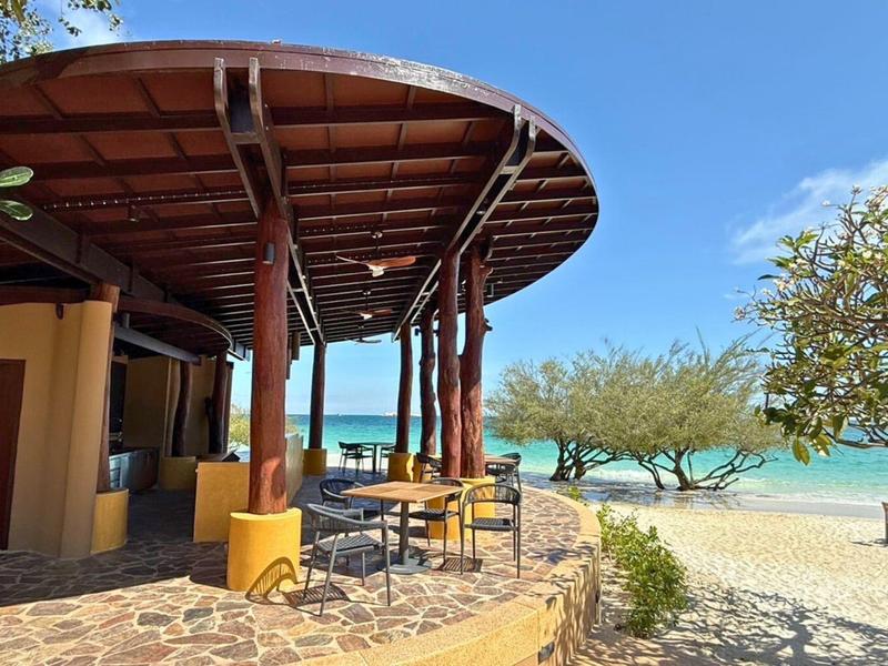 Open beach pavilion with tables and chairs beside the sandy shore and sea.
