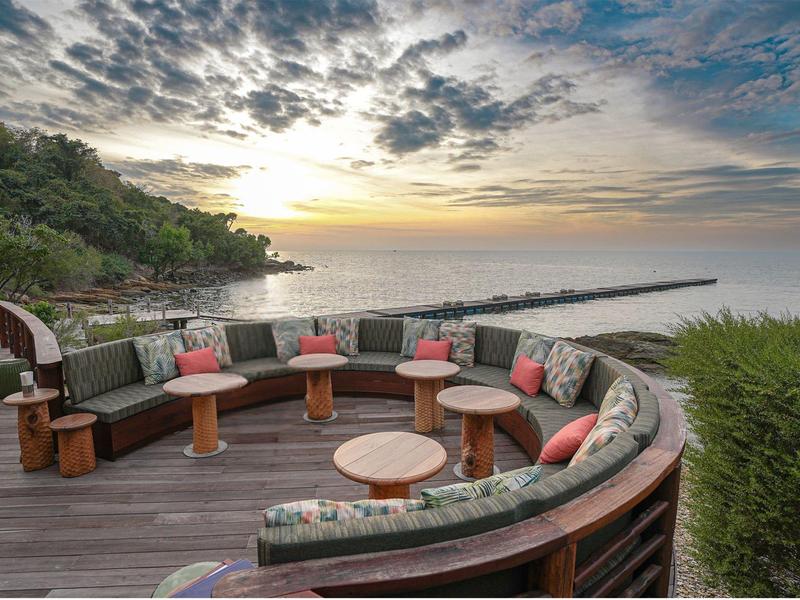Round wooden terrace with cushioned seats and tables overlooking the sea at sunset.