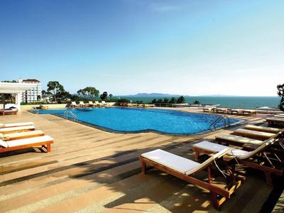 Großer Pool mit Holzliegen auf sonniger Terrasse, dahinter Meer und blauer Himmel.