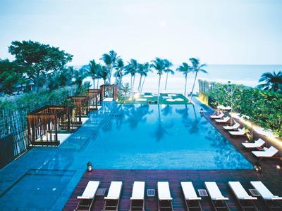 Infinity-zwembad met ligstoelen en palmbomen bij zee onder een heldere lucht.